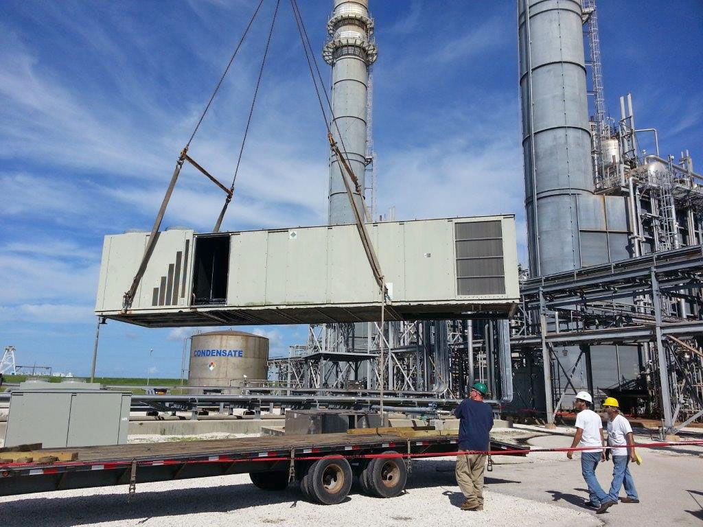 Workers guide a large air handling unit hanging from rigging straps at an industrial plant