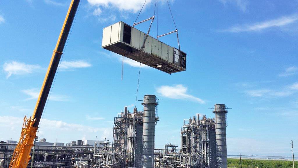A crane lifts a large commercial air handling unit into place above an industrial plant